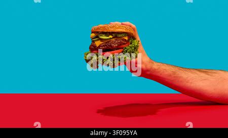 Hand holding large cheeseburger on turquoise and red studio background. Stock Photo