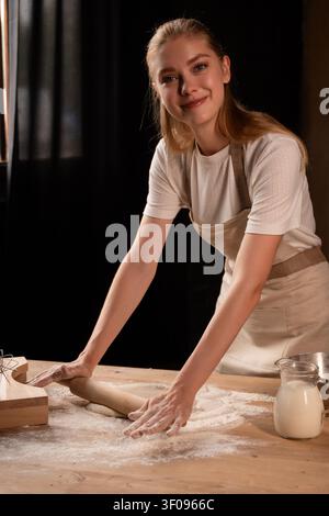Woman baking homemade dough with rolling pin and flour, preparing bread while smiling in a dark kitchen Stock Photo