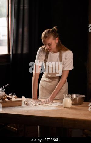 Woman wearing apron kneading dough with flour on wooden table preparing homemade pastry in kitchen Stock Photo