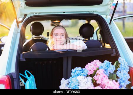 Golden hour portrait of a young woman enjoying nature, travel and leisure beside a stylish pastel vehicle and blooming hydrangeas Stock Photo