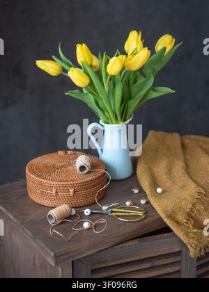 Elegant bouquet of vibrant yellow tulips in a light blue enamel vase, placed on a dark rustic wooden table with a golden scarf, scissors, and string. Stock Photo