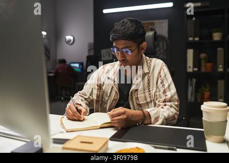 Asian Gen Z Man Taking Notes at Desk during Evening Study Stock Photo