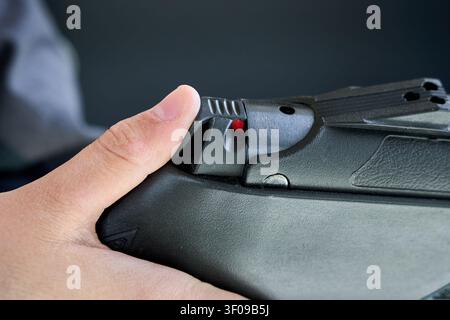 Close-up view shows young man thumb engaging manual safety switch on black firearm. Red dot indicator visible, signifying weapon is ready to fire. Sha Stock Photo