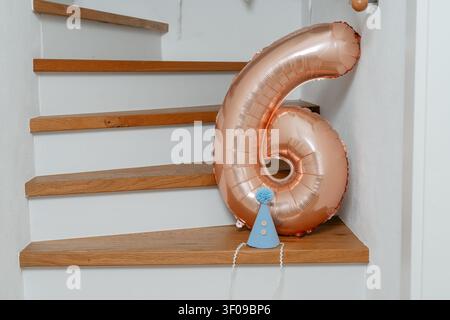 A number six balloon rests on a wooden staircase, adorned with a blue party hat, celebrating a birthday party in an indoor setting with white walls Stock Photo