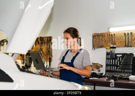 Woman car mechanic performing car diagnostics using a laptop beside a vehicle with open hood Stock Photo
