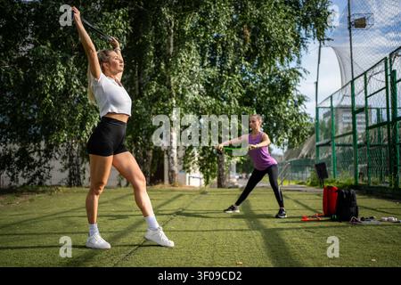Outdoor fitness training and flexibility workout. Two sportswomen doing resistance band stretching exercises on sports field on summer day. Stock Photo
