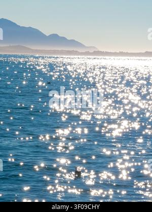 Bright sun reflection creating a sparkling bokeh effect on the blue ocean surface with coastal mountains in the background, Kaikoura, New Zealand Stock Photo