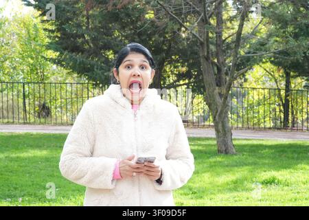 Young woman outdoors, surprised expression. She holds smartphone, dressed in white fleece. Green grass, trees, and fence form the background. Natural Stock Photo