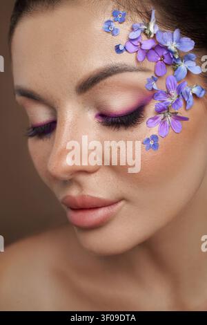 Beauty  woman poses with makeup featuring purple flowers on one side of her face. She has dark hair and makeup that highlights her eyes. The scene is Stock Photo