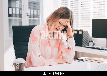 Young professional woman suffering from headache while analyzing financial charts and graphs on computer, experiencing work related stress in modern o Stock Photo