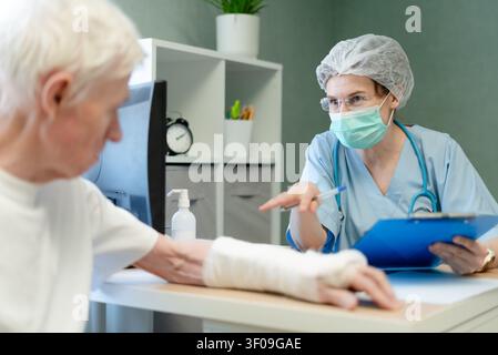 Doctor pointing at patient cast during hands-on medical examination. Professional physician checking elderly man arm injury in clinic. Attentive healthcare worker demonstrating care through assessment Stock Photo