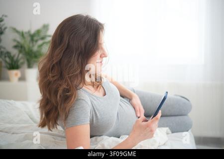 A pregnant woman rests on a bed on a video call in a bright bedroom. The expectant mother using a mobile phone presents a connected, relaxed moment of comfortable communication at home. Stock Photo