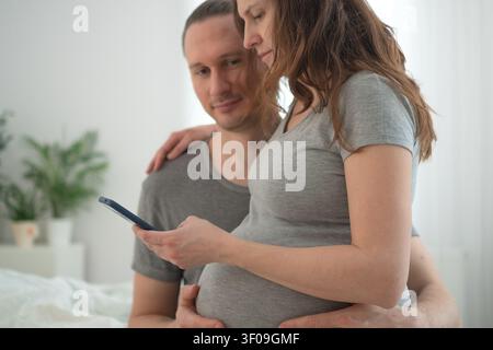 Pregnant woman and husband standing together in bright bedroom sharing pregnancy news on a phone call. Expecting mother with arm around waist while partner holds phone communicating with family. Stock Photo