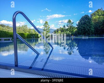 A scenic view of a crystal clear outdoor public swimming pool on a bright sunny day. A metallic steel handrail leads into the calm blue water, which p Stock Photo