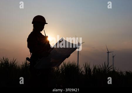 Female engineer in protective gear examining construction blueprints outdoors against a sunset backdrop featuring sustainable wind energy infrastructu Stock Photo