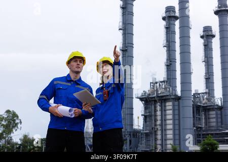 Professional engineers in safety gear inspect an energy facility reviewing infrastructure plans on a tablet to coordinate maintenance operations. Stock Photo