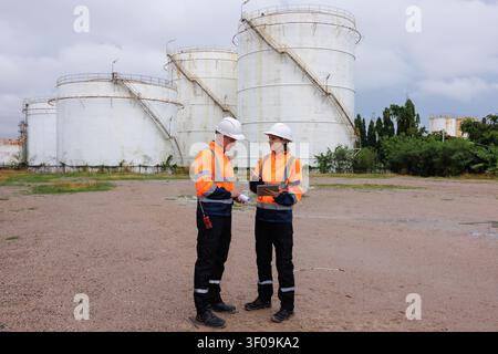 Two engineers in highvisibility safety clothing review technical data on a tablet before large storage tanks at a petrochemical site. Stock Photo