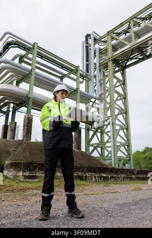 Female professional in protective gear using a tablet to monitor industrial piping and infrastructure in a utility plant setting. Stock Photo
