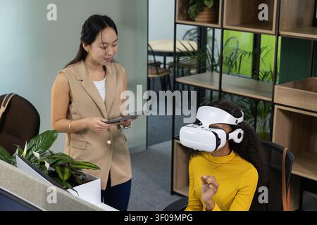Diverse female coworkers testing VR headset while monitoring on tablet at office, copy space. Virtual reality, collaboration, device, modern, technology, teamwork, greenery Stock Photo
