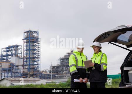 Two engineers in hard hats and reflective safety jackets consult digital plans on a tablet at an industrial plant site. Stock Photo