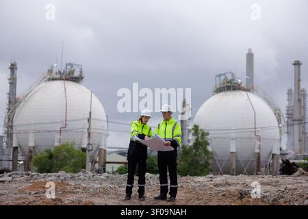 Two industrial engineers wearing safety helmets and reflective jackets collaborate on blueprints at a refinery with gas storage tanks in the backgroun Stock Photo