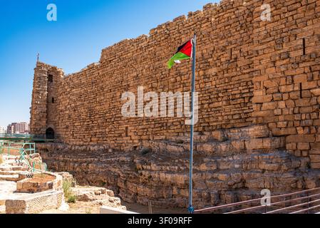 A tall and imposing squared stone wall of the Al-Karak Castle dominates the daytime scene. In the foreground, the flag of Jordan waves on a metallic p Stock Photo