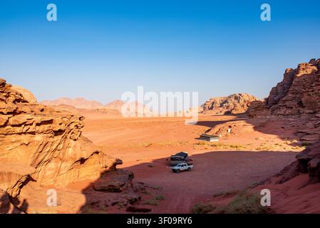 A white pick-up truck stands next to dark Bedouin tents inside a sandy canyon in Wadi Rum. Majestic sandstone rock walls enclose the orange valley, il Stock Photo