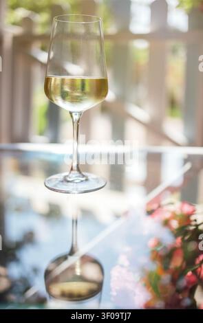 Glass of white wine positioned on a reflective table, surrounded by a blurred garden setting with colorful flowers in the background Stock Photo