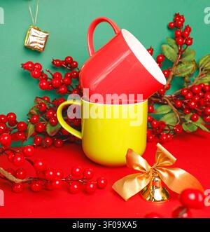Christmas mockup red and yellow ceramic mugs stacked on red table against green background. New year bright cups template with winter holly berries Stock Photo