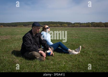 Couple enjoying fresh air while sitting on green grass in a vast open field with trees in the background, a woman looking up at the sky and a man hold Stock Photo