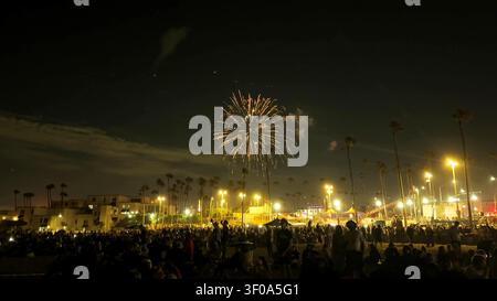 A bright golden firework explodes in the dark night sky over a crowded beach with palm trees. Stock Photo