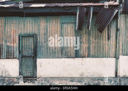 A front-facing exterior shot of a run-down structure featuring rusted corrugated iron panels, a broken roof overhang, an old screen door, and a weathe Stock Photo