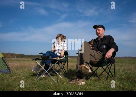 Young couple camping in a sunny meadow, sitting by a tent and portable grill, relaxing together under blue skies while enjoying outdoor cooking, bondi Stock Photo