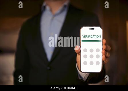 Man dressed in a black suit presents a smartphone showing a verification screen with circular input fields, background appears blurred and indoor Stock Photo