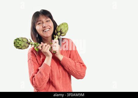 Woman smiles brightly while holding two artichokes. She wears a warm, coral-colored knit sweater. Her dark hair frames her cheerful expression. White Stock Photo