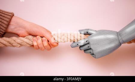 A human hand and a robotic hand hold a thick rope, set against a soft pink background, illustrating the interaction between human and artificial intel Stock Photo