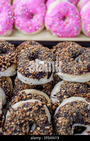 Close up of chocolate peanut glazed donuts and pink frosted pastries stacked on a bakery display counter at a Thai street food night market. Stock Photo