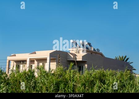 Two-story beige residential house with solar panels and water heating systems on the rooftop. Green reed plants in foreground and clear blue sky backg Stock Photo