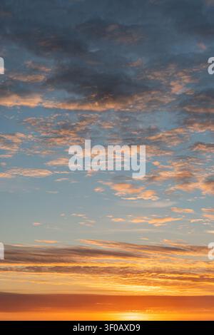 Sunset scene with scattered clouds, orange, yellow, and blue hues, horizon line, varying cloud shapes and sizes, warm glow, serene transition from day Stock Photo
