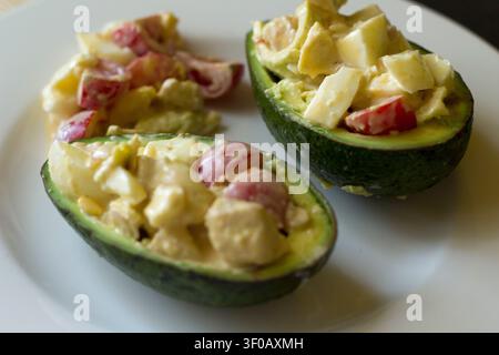 Two avocado halves stuffed with chopped boiled egg, cherry tomatoes and creamy dressing on a white plate, close up Stock Photo