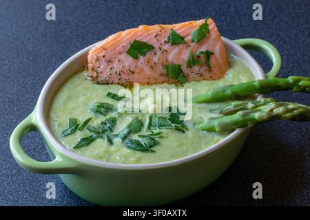 Creamy asparagus soup topped with salmon fillet, dill and parsley in a green casserole, asparagus tips on the right, close angle, dark surface Stock Photo