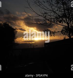 A beautiful scenic landscape shot at dusk, capturing the deep golden and orange glow of the setting sun breaking through dark clouds behind a silhouet Stock Photo