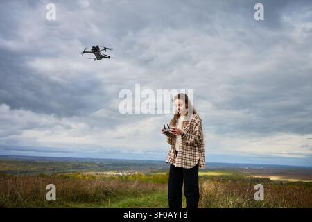 Woman controlling drone flying in cloudy sky over scenic landscape. Female pilot using remote controller on hill, aerial photography and exploration concept of technology Stock Photo