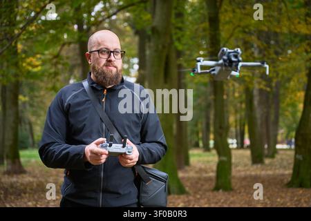 Man controls drone while watching it fly nearby. Bearded man operating quadcopter in park, focused on flight control during outdoor session, concept of modern technology and hobby Stock Photo