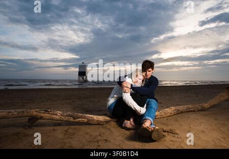 Young couple sitting on drift wood log hugging and looking out to sea on beach at sunset Stock Photo