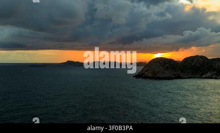 Golden sunlight pierces through heavy dark storm clouds over rocky islets in the sea at sunset in Lindesnes, Norway Stock Photo