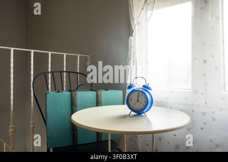 Blue alarm clock on a white table with turquoise suitcase, black chair and star curtain, cozy rural house bedroom prepared for travel. Stock Photo