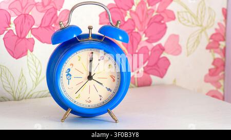 Blue vintage alarm clock on a white table with pink floral wallpaper and soft light, simple still life about time, travel and rural accommodation. Stock Photo