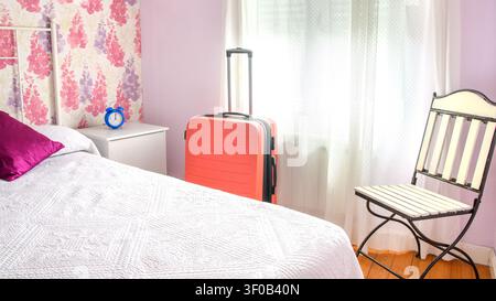 Salmon carry-on suitcase beside a white bed, vintage chair, blue alarm clock and sheer curtains in a bright rural house bedroom. Stock Photo