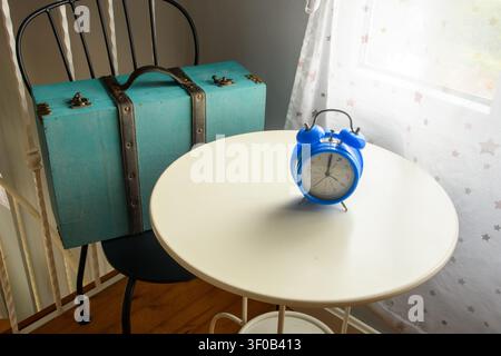 Blue alarm clock on a round white table beside a turquoise vintage suitcase and star curtain, evoking travel timing in a rural house. Stock Photo